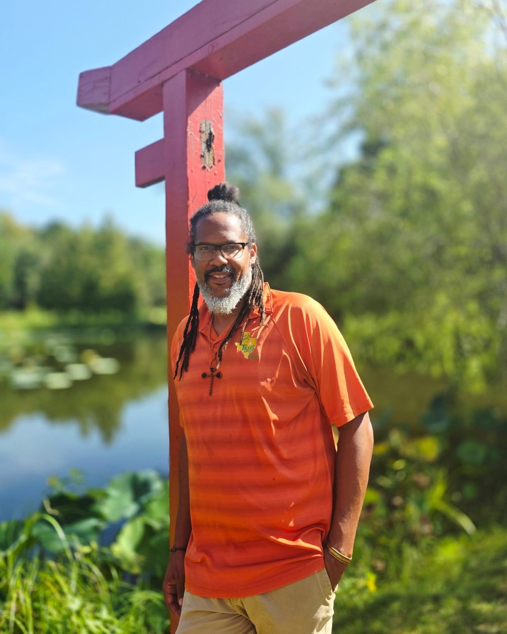 Chef Tony Tejas Ingram in an orange Tony's Tejas Salsa shirt, standing beside a red wooden gate by a pond