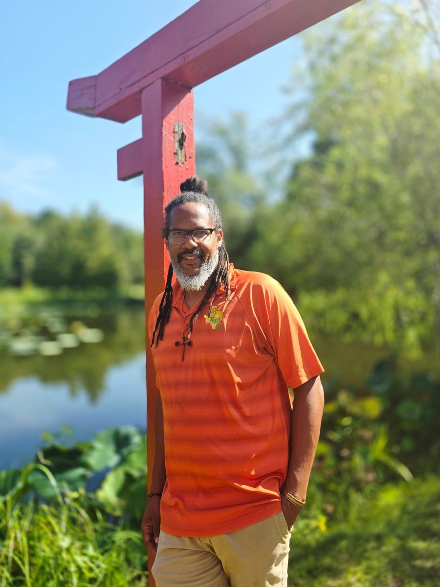 Chef Tony Tejas Ingram in an orange Tony's Tejas Salsa shirt, standing beside a red wooden gate by a pond